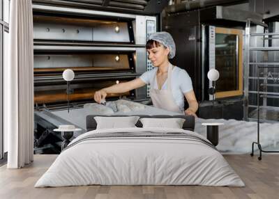 Young caucasian woman baker is preparing bread dough and cut it with knife for bread baker process in electric oven at baking manufacture factory Wall mural