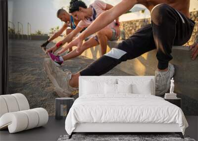 A group of three people, two men and a woman, do some stretching exercises, they are sitting and touch their feet with their hand, warm-up pre work out in the open air in an outdoor gym. Wall mural