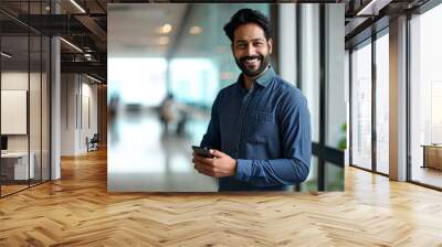 Happy Indian business man holding mobile cell phone device looking at camera standing at work. Smiling professional businessman using finance banking apps smartphone working on cellphone,Generative AI Wall mural