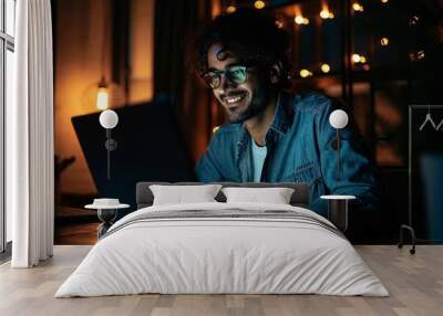 Smiling young curly indian latin ethnic business man or student wearing glasses remote working overtime, learning online late at night at home or in dark office using laptop computer, Generative AI Wall mural