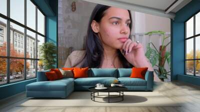 Beautiful Indian woman in a thinking pose sitting near the plants at the apartment Wall mural