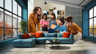 Colorful portrait of young female teacher talking to diverse group of kids using computer during class in school library Wall mural