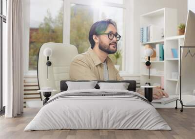 Content creative young Asian man with beard sitting at desk and typing on keyboards while communicating with client online Wall mural