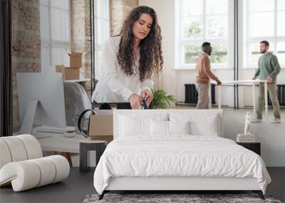 Curly-haired young woman in white blouse standing at wooden desk with computer and unpacking personal stuff from box in new office Wall mural