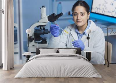 Portrait of content young Indian female pharmacist in lab coat and surgical gloves sitting at desk and mixing reagents in test tube using pipette Wall mural