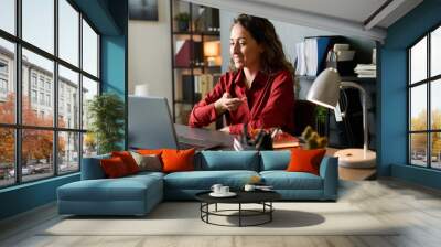 Smiling woman sitting at desk gesturing while working on laptop in well-organized, modern office space with shelves, books, plants, and warm lighting Wall mural