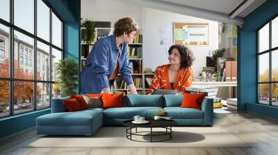 Two women having discussion in office with shelves, books, and plants around. One woman standing and gesturing, other woman sitting and holding tablet, both engaged in conversation Wall mural