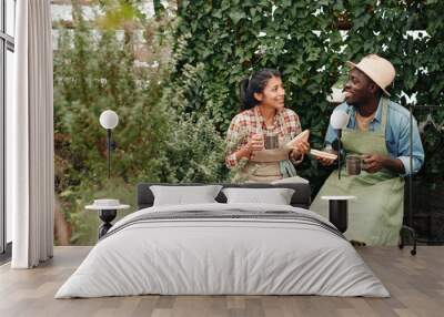 Two young ethnically diverse co-workers wearing aprons enjoying having lunch together in modern greenhouse Wall mural