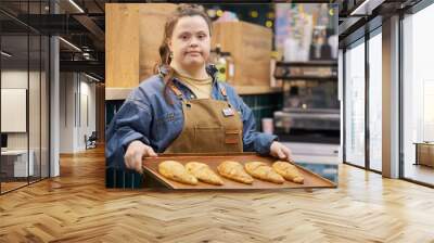 Waist up portrait of confident young woman with Down syndrome working in bakery and looking at camera holding tray with fresh pastry copy space Wall mural