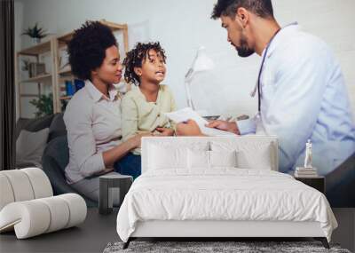 African American male pediatrician with stethoscope talking to little girl sitting on mother lap. Wall mural