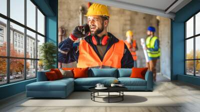 Construction site manager standing  wearing safety vest and helmet, talking to the phone at construction site. Wall mural