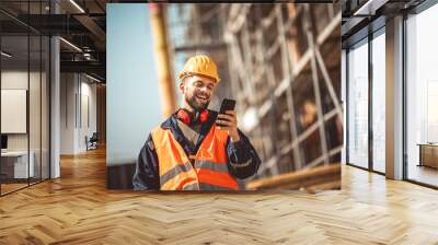 Construction site manager standing  wearing safety vest and helmet,using his phone at construction site. Wall mural