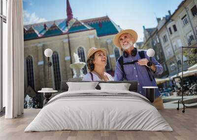 Elderly married couple exploring new city. They are wearing hats and backpacks. Wall mural