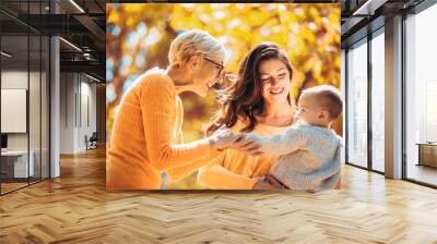 Grandmother and mother smiling at baby in autumn park. Wall mural