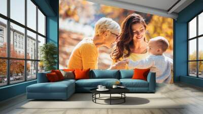 Grandmother and mother smiling at baby in autumn park. Wall mural