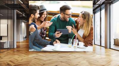 Group of four friends having a coffee together. Two women and two men at cafe talking laughing and enjoying their time using digital tablet. Wall mural