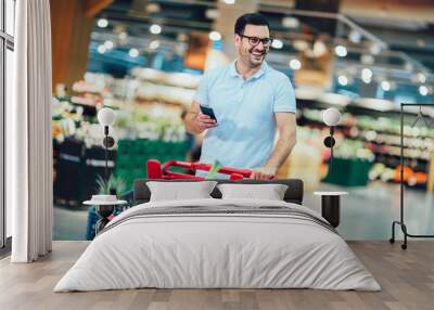 Handsome man shopping in supermarket pushing trolley and holding phone. Wall mural