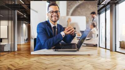 Happy businessman using laptop with team discussing project in the background. Wall mural