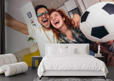 Happy young couple spending weekend together at home sitting on sofa watching soccer play live on tv and cheering for favourite team Wall mural