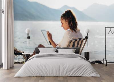 Smiling woman sitting on deck chair by the sea using tablet on a sunny day Wall mural