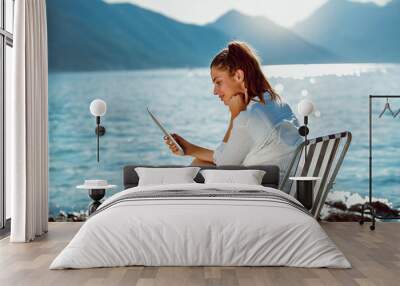 Smiling woman sitting on deck chair by the sea using tablet on a sunny day Wall mural