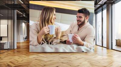 Smiling young couple dressed in bathrobes lying on bed and drinking coffee Wall mural