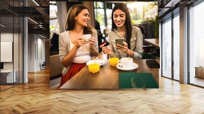 Two girl best friends met at the cafe to shop online together. They are using mobile phone and credit card. Friends hang out and drink coffee and orange juice. Wall mural