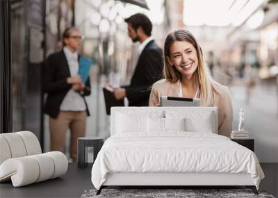 Young happy businesswoman holding digital tablet outside of modern building, businesspeople in background. Wall mural