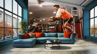 Young man doing exercise on elliptical cross trainer in sport fitness gym club Wall mural