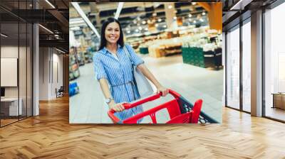 Young woman shopping in supermarket pushing trolley and smilling. Wall mural