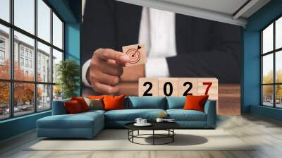 Business planning and strategy in 2027. Hand businessman holding wooden cubes with dartboard icon and wooden cubes with the letters 2027 on a table Wall mural