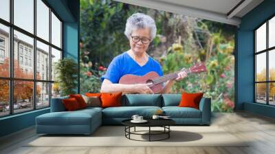 Portrait of elderly woman playing ukulele in her garden. Relaxing by singing and play small guitar happy and enjoy life after retired. Concept of old people and health care. Space for text Wall mural