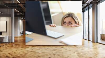 Scared female manager hidden behind office desk and looking at computer screen. Wall mural