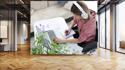 Woman in wheelchair sits at work desk wearing headphones and makes notes in notebook Wall mural