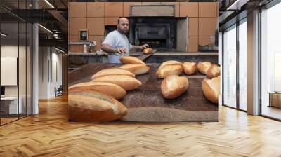 Baker. A young handsome bakery worker on the background of bread, takes bread from a stove with a wooden shovel. Industrial production of bakery products. a man in the baker's special clothing. Bakery Wall mural