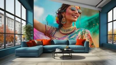 A beautiful young woman celebrates Holi, the festival of colors, in a traditional Indian saree and jewelry. She playfully throws colors while posing fashionably with sunglasses. Wall mural