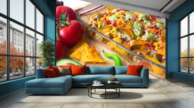 Baked nacho dish in a glass baking pan with corn tortilla chip, pico de gallo salsa, avocado guacamole chipotle, beans, jalapenos and Monterrey jack cheese on a wooden board surrounded by ingredients Wall mural