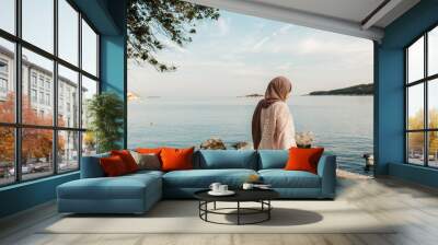 portrait of young European Muslim women with hijab sitting on the beach with hat in her hand. She is happy and relaxed. Sea is in the background. She is looking in the distance. Wall mural