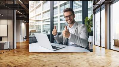 happy businessman in eyeglasses showing thumbs up while working with laptop in office Wall mural