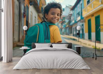 A Latino boy wearing a green backpack smiles as he stands on the street and turns to look at the camera. It's a typical street in a Spanish-American city. Back to school. Wall mural