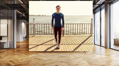 Healthy young male dressed in sportswear poses on pier. Sportsman walking by the sea at sunset. Photo of handsome athletic young man walking towards camera Wall mural