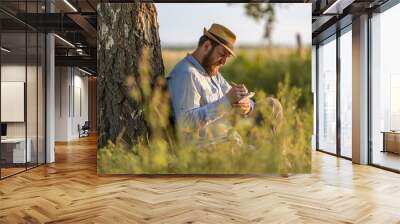 an adult man in a hat with a beard writes poetry or prose sitting by a tree Wall mural