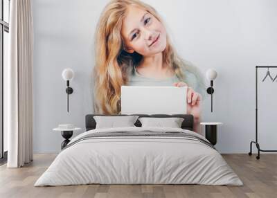 Beautiful girl with blank canvas board for message or photo. Copy space. Cute teen caucasian girl with blond hair holding in her hands mockup poster and standing against grey wall. Wall mural