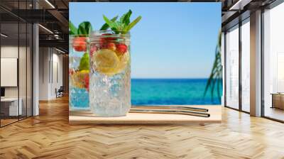 Summer cocktails, beach bar concept. Two drinking glasses of hard seltzer cocktail with strawberry, lemon and mint, beautiful sea on background. Summer resort concept. Wall mural