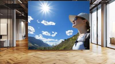 A hiker wearing a sun hat and sunglasses, looking out over a scenic vista with a bright sky above. Wall mural