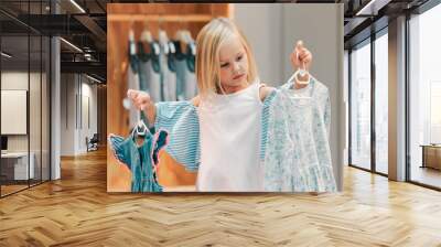 Child, fashion and shopping while choosing kids clothing in a shop, store or boutique with a cute girl holding dresses thinking or picking color choice. Little kid shopper or customer buying clothes Wall mural