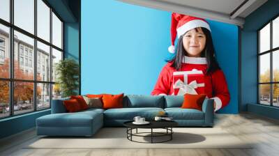 Christmas during coronavirus covid19.Little asian santa girl holding gift box isolated on blue background.Christmas winter celebration at home.Social distancing, Christmas present surprise gift. Wall mural