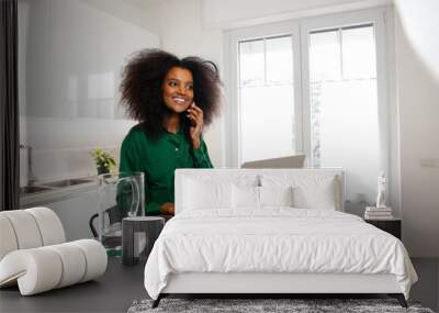 An africano woman cheerfully calling over the phone at home while working with her laptop and water beside her in her home office Wall mural