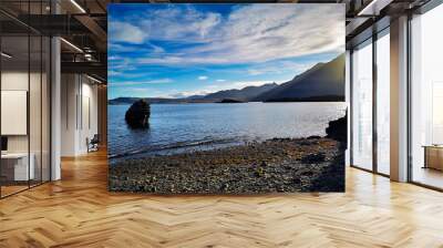 A peaceful Icelandic coastline with a rocky beach, calm waters, and distant mountains under a bright blue sky. A large rock formation stands near the shore, adding to the scenic beauty Wall mural