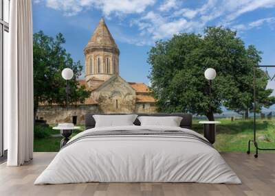 Church of Khirsa monastery of Saint Stephen. Stone walls, orange tile roof, green lawn, blue bright sky with clouds, gravestones and huge tree. Visible Alazani valley Wall mural